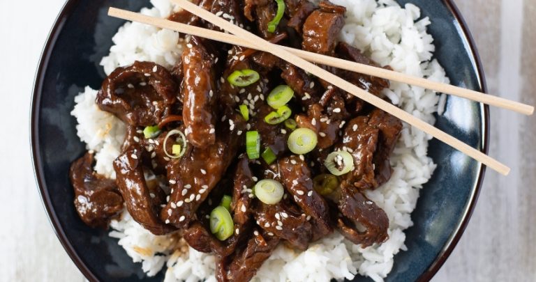 a dark blue bowl filled with tender Crock Pot Mongolian Beef served over a bed of cooked white rice with sliced green onions as the garnish.